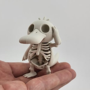 Può includere: Statuetta beige chiaro di un'anatra con un design a scheletro. L'anatra ha un becco grande, orbite rotonde e una piccola coda. Lo scheletro è visibile attraverso il corpo dell'anatra. La statuetta è tenuta in una mano.
