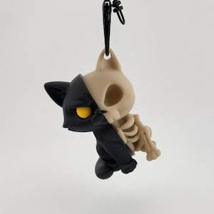 Peut inclure: Figurine de chat noir et beige avec un design squelette d'un côté, attachée à un porte-clés noir. Le chat a des yeux jaunes. Une figurine de chat bicolore avec une touche effrayante.