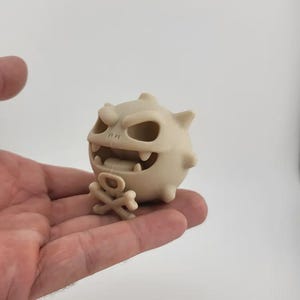 May include: A beige, cartoonish figurine of a monster with a skull-like face, sharp teeth, and a small crossbones. The figurine is held in a person's hand against a white background. The monster has a round body with small spikes.