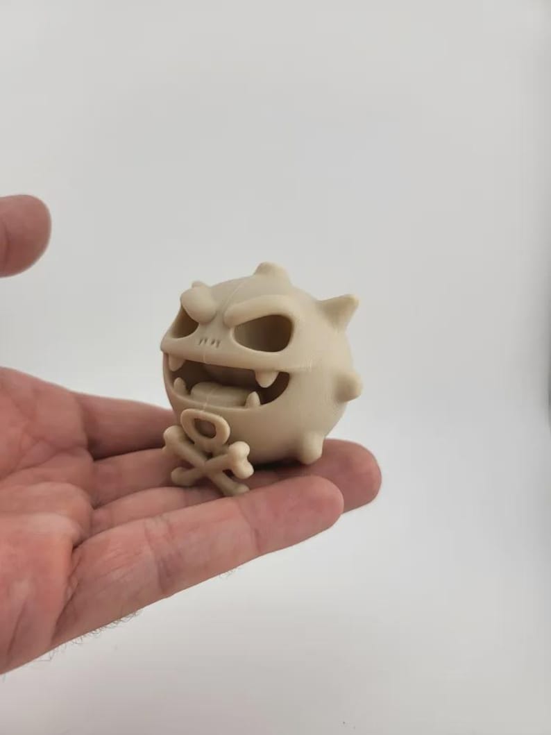 May include: A beige, cartoonish figurine of a monster with a skull-like face, sharp teeth, and a small crossbones. The figurine is held in a person's hand against a white background. The monster has a round body with small spikes.