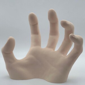 May include: A pale pink ceramic hand sculpture with an open palm and outstretched fingers. The hand is positioned as if reaching out or grasping.
