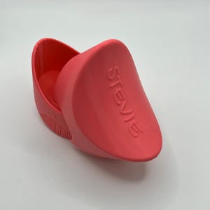 K&ouml;nnte beinhalten: Ein korallenfarbener Schuhanzieher mit dem Namen "STEVIE" auf der Seite. Der Schuhanzieher hat ein gebogenes Design mit einem kleinen, geriffelten Abschnitt an der Basis. Das Produkt besteht aus einem festen Material.