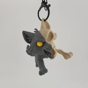 Peut inclure: Un porte-clés bicolore représentant un loup stylisé. Un côté est gris, l'autre est un crâne beige. Le loup a des yeux jaunes et est attaché à un fermoir noir.