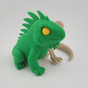 Peut inclure: Figurine d'iguane vert avec des yeux jaunes et un squelette beige. L'iguane est l'élément principal, avec une surface texturée détaillée. Le squelette est positionné derrière l'iguane. La figurine est sur une surface blanche.