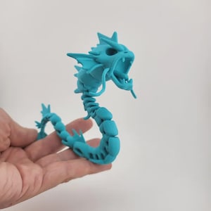 Puede incluir: Figura articulada de drag&oacute;n turquesa con boca abierta y p&uacute;as. El cuerpo del drag&oacute;n est&aacute; segmentado, lo que permite posarlo. La figura se sostiene en una mano sobre un fondo blanco liso. El drag&oacute;n es una criatura de fantas&iacute;a.