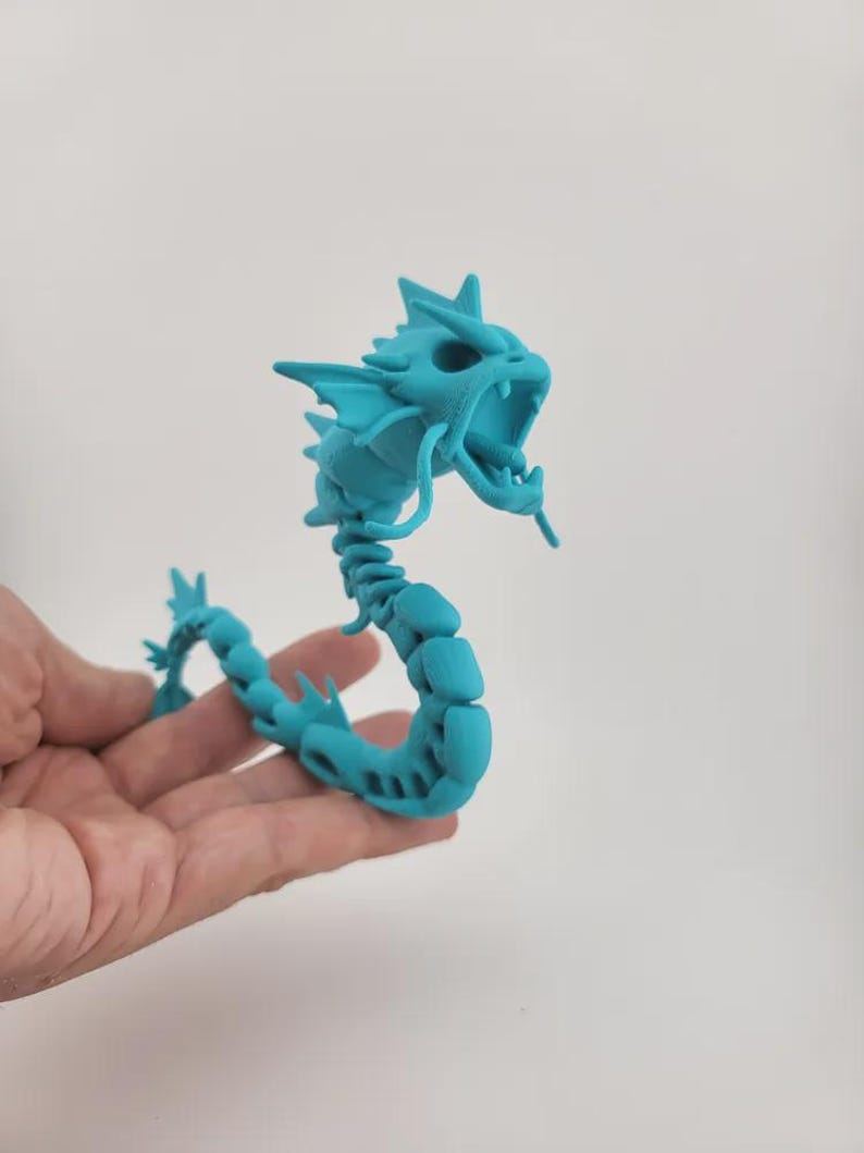 Puede incluir: Figura articulada de drag&oacute;n turquesa con boca abierta y p&uacute;as. El cuerpo del drag&oacute;n est&aacute; segmentado, lo que permite posarlo. La figura se sostiene en una mano sobre un fondo blanco liso. El drag&oacute;n es una criatura de fantas&iacute;a.