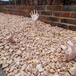 May include: Three realistic-looking human hands are sticking out of a bed of small, light brown gravel. The hands are posed as if reaching up from the ground.