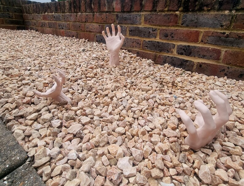 May include: Three realistic-looking human hands are sticking out of a bed of small, light brown gravel. The hands are posed as if reaching up from the ground.