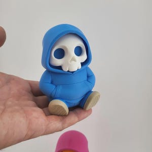 May include: A small, blue figurine of a skull wearing a hoodie. The skull is white with large eye sockets and a small nose. The hoodie is a bright blue, and the figurine is sitting with its hands in its pockets. The feet are tan.