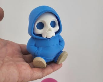 May include: A small, blue figurine of a skull wearing a hoodie. The skull is white with large eye sockets and a small nose. The hoodie is a bright blue, and the figurine is sitting with its hands in its pockets. The feet are tan.