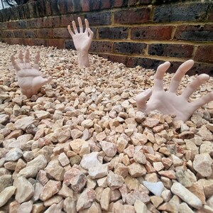 May include: Two hands reaching up from a pile of tan gravel stones. The hands are pale and appear to be made of plastic.