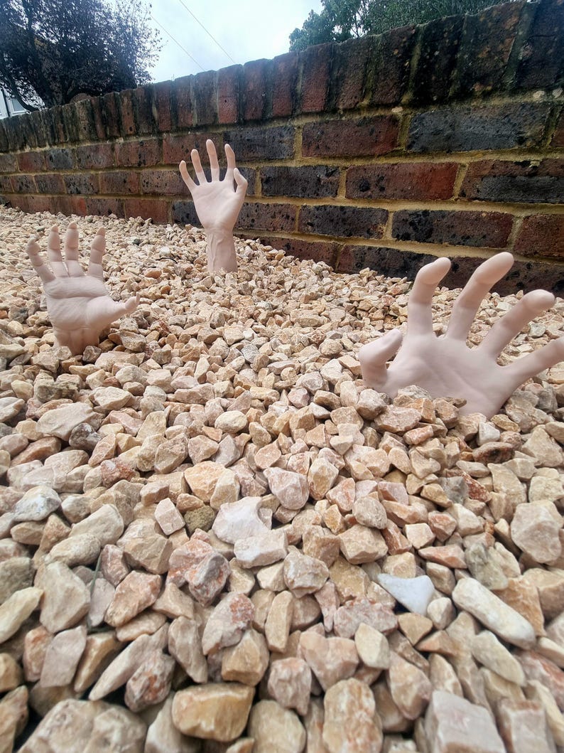 May include: Two hands reaching up from a pile of tan gravel stones. The hands are pale and appear to be made of plastic.