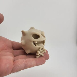 May include: A beige, cartoonish figurine of a round creature with a menacing expression. It has large teeth, a wide open mouth, and a small skull and crossbones at its base. The figurine has several small spikes.