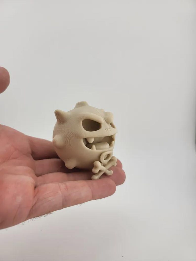 May include: A beige, cartoonish figurine of a round creature with a menacing expression. It has large teeth, a wide open mouth, and a small skull and crossbones at its base. The figurine has several small spikes.