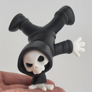May include: A whimsical figurine of the Grim Reaper in a handstand pose. The figure is primarily black with a white skull face and hands. The Reaper's outfit includes a hooded cloak and boots.