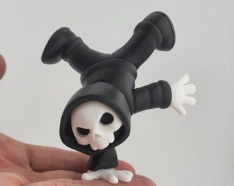May include: A novelty figurine of the Grim Reaper performing a handstand. The figure is predominantly black with a white skull face and hands. The Reaper is wearing a hooded cloak and boots.