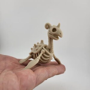 May include: A beige, skeletal figurine of a mythical creature, possibly a dragon or dinosaur, with a long neck, horns, and a detailed rib cage. The figurine is held in a person's hand, showcasing its size and intricate design.