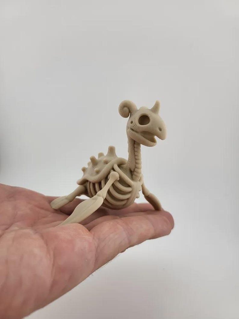 May include: A beige, skeletal figurine of a mythical creature, possibly a dragon or dinosaur, with a long neck, horns, and a detailed rib cage. The figurine is held in a person's hand, showcasing its size and intricate design.