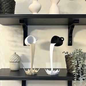 May include: Two white ceramic cups with black and white paint pouring out of them, forming a splash shape. The cups are on a black shelf with other white ceramic sculptures.