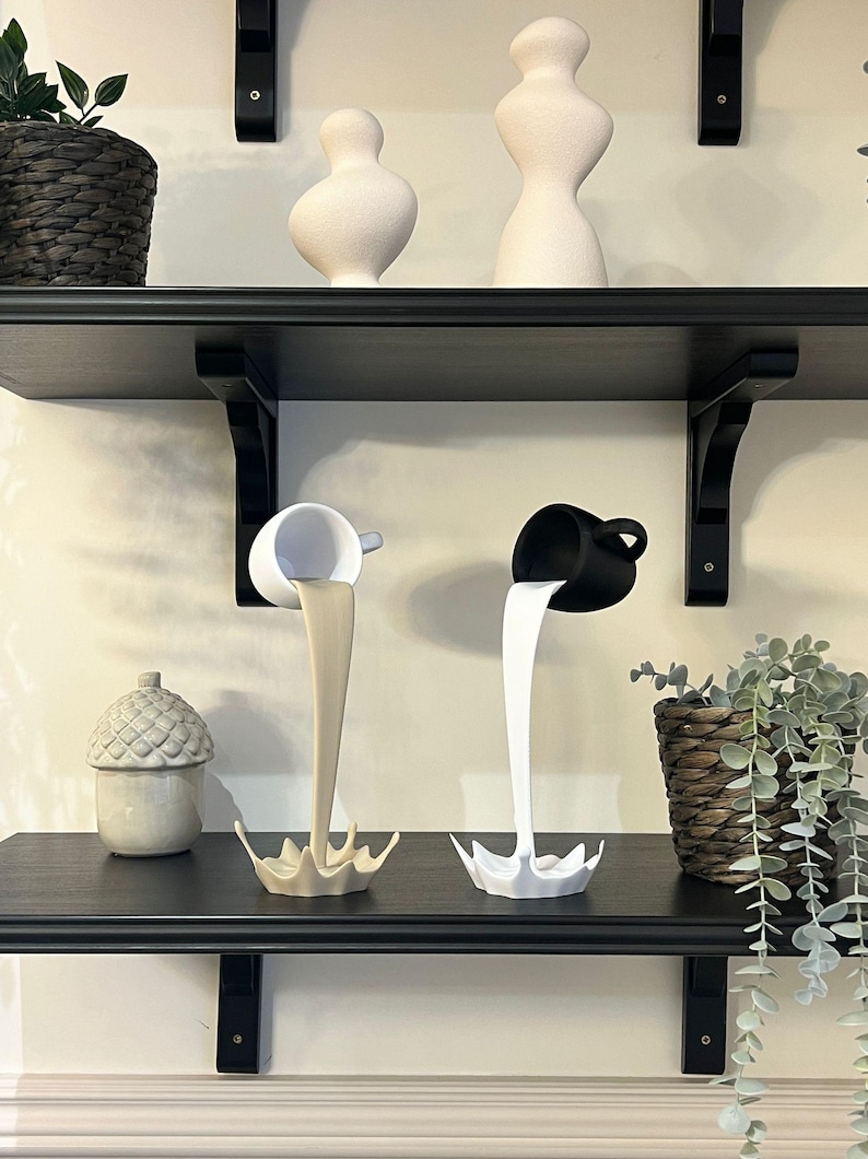 May include: Two white ceramic cups with black and white paint pouring out of them, forming a splash shape. The cups are on a black shelf with other white ceramic sculptures.