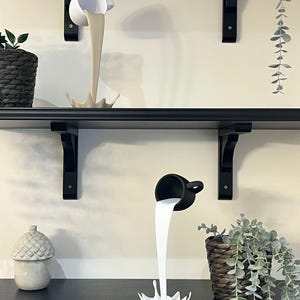 May include: Two ceramic cups pouring out a liquid, one white and one black, on a black shelf with a plant in a wicker pot.