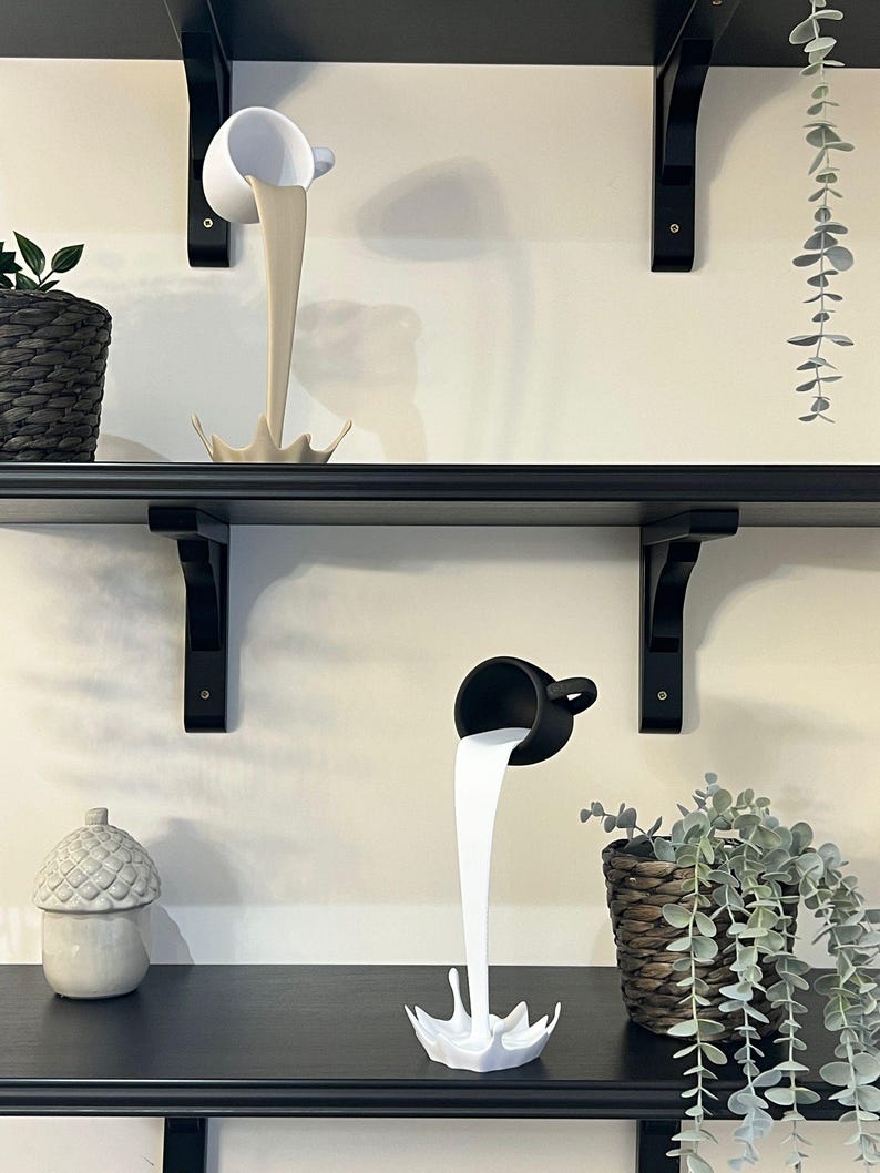 May include: Two ceramic cups pouring out a liquid, one white and one black, on a black shelf with a plant in a wicker pot.
