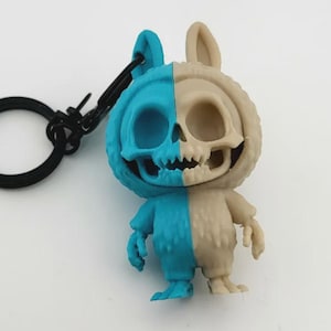 Peut inclure: Un porte-clés bicolore représentant une figurine de lapin-squelette. La figurine est divisée verticalement, avec un côté turquoise et l'autre beige. Le porte-clés a un anneau et un fermoir noirs.