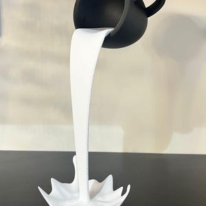 May include: A black ceramic cup pouring white liquid into a white ceramic splash. The cup is upside down and the liquid is frozen in mid-air.