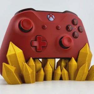 May include: A red Xbox controller on a yellow crystal base. The controller features the Xbox logo and buttons. The base is composed of multiple yellow, pointed crystal shapes, creating a decorative stand.
