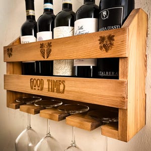 Może przedstawiać: Drewniany stojak na wino z pięcioma butelkami wina i czterema kieliszkami do wina. Stojak ma półkę na butelki i dolną półkę na kieliszki. Stojak jest oznaczony napisem "Good Times".