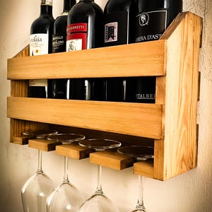 Może przedstawiać: Drewniany stojak na wino montowany na ścianie z pięcioma butelkami czerwonego wina i czterema kieliszkami do wina wiszącymi poniżej. Stojak ma dwie półki i jest wykonany z jasnobrązowego drewna.