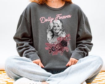 Könnte beinhalten: Ein anthrazitfarbenes Sweatshirt mit Rundhalsausschnitt und einem schwarz-weißen Porträt von Dolly Parton auf der Brust. Über dem Porträt steht der Text Dolly Forever in neonpinker Schrift. Unter dem Porträt befinden sich dunkelrosa Blumenmotive.