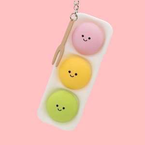 Puede incluir: Un llavero caprichoso con tres macarons sonrientes en una bandeja blanca. Los macarons son rosas, amarillos y verdes, con un tenedor pequeño de madera y una cadena plateada. El fondo es rosa suave.