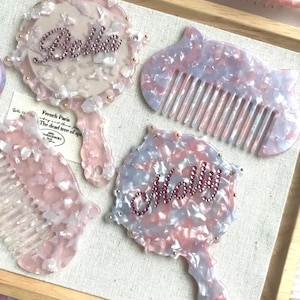Op de afbeelding: Een verzameling roze, witte en blauw gemarmerde haar kammen en handspiegels. Twee spiegels zijn versierd met de namen "Belle" en "Molly" in roze strass-steentjes. De items zijn gerangschikt op een houten dienblad.