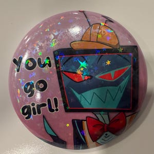 Puede incluir: Un botón redondo rosa con purpurina holográfica. El botón presenta un personaje de dibujos animados con una cara de pantalla, una pajarita y el texto "You go girl!". El personaje tiene un sombrero marrón y una expresión amenazante.