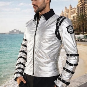May include: A metallic silver long-sleeve jacket featuring a black collar, a full-length front zipper, and black buckled straps encircling both sleeves. A black harness-style strap crosses the left shoulder, adorned with a circular black patch.