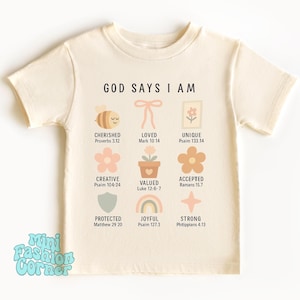 Op de afbeelding: Een beige T-shirt met de tekst "GOD SAYS I AM" in zwart gedrukt. Het shirt heeft verschillende kleurrijke illustraties en bijbelteksten, waaronder "Gekoesterd, Spreuken 3:12", "Geliefd, Markus 10:14", "Uniek, Psalm 133:14", "Creatief, Psalm 104:24", "Waardevol, Lukas 12:6-7", "Aanvaard, Romeinen 15:7", "Beschermd, Mattheüs 29:20", "Vrolijk, Psalm 127:3", en "Sterk, Filippenzen 4:13".