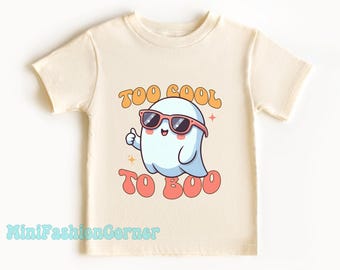 Può includere: Una maglietta color crema con un fantasma dei cartoni animati che indossa occhiali da sole e il testo "Too Cool To Boo" in un font retrò.