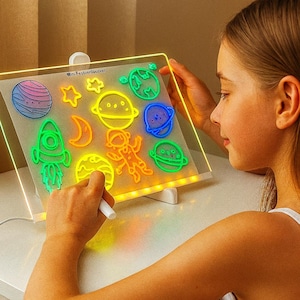 May include: A light-up drawing board with colorful space-themed shapes. The board features a variety of glowing planets, stars, and a rocket ship. A child is using a white marker to draw on the board. The text "Mini Fashionlover" is visible.