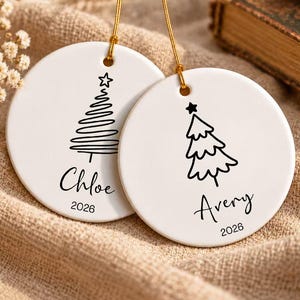 May include: Two round white ceramic ornaments with gold hanging strings. One ornament features a line-art Christmas tree, the name Chloe, and the year 2026. The other ornament features a different line-art tree, the name Avery, and the year 2026.