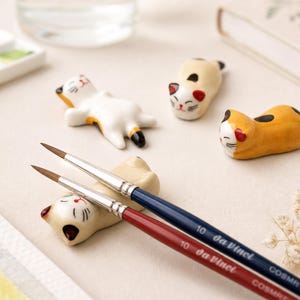 May include: Four ceramic cat-shaped rests in white, cream, and orange with black spots, paired with two paintbrushes. The brushes feature silver ferrules, dark blue and red wooden handles, and are marked with the text 10 Da Vinci COSMOTOP.