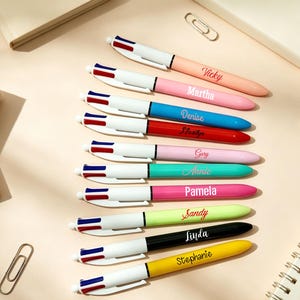 May include: A collection of ten multi-color ballpoint pens arranged in a vertical row. Each pen features a white top section with blue and red buttons, and a solid-colored barrel personalized with a name in white or black script lettering.