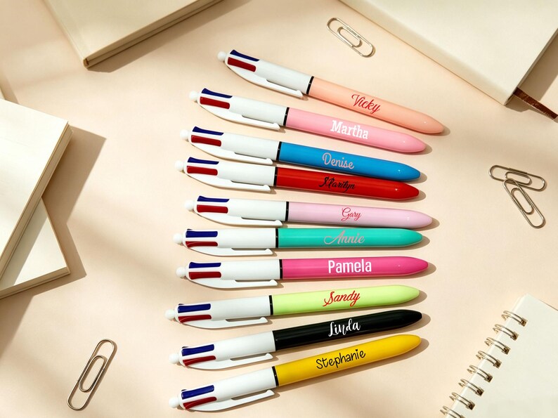 May include: A collection of ten multi-color ballpoint pens arranged in a vertical row. Each pen features a white top section with blue and red buttons, and a solid-colored barrel personalized with a name in white or black script lettering.