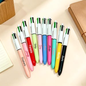 May include: A collection of nine multi-color ballpoint pens arranged in a fan shape. Each pen features a white barrel with a green and black top mechanism, and a unique name printed in cursive script on the barrel, including Stephanie, Pamela, Vicky, Gary, Annie, Marilyn, Denise, Martha, and Linda.