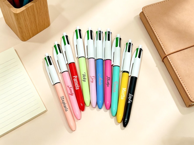 May include: A collection of nine multi-color ballpoint pens arranged in a fan shape. Each pen features a white barrel with a green and black top mechanism, and a unique name printed in cursive script on the barrel, including Stephanie, Pamela, Vicky, Gary, Annie, Marilyn, Denise, Martha, and Linda.