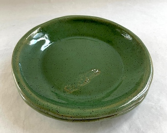 May include: Three handmade ceramic plates stacked together. The plates are a deep green with a speckled texture and a glossy finish. The edges are slightly irregular, giving them a rustic look. The plates are stacked on top of each other, showing their handmade quality.