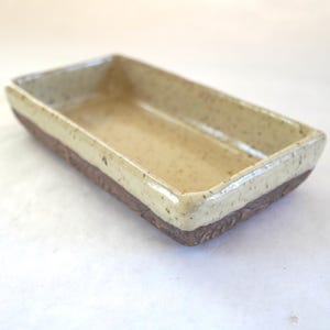 May include: A rectangular ceramic dish with a speckled beige interior and a brown base. The rim is a lighter beige colour. The base has a textured, carved design. The dish is likely handmade and suitable for serving or display.