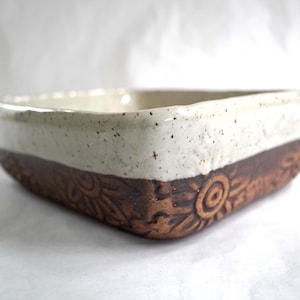 May include: A square ceramic bowl featuring a two-tone design. The upper portion is finished in a speckled off-white glaze, while the lower portion displays a dark brown clay body with an embossed floral pattern featuring circular centers.
