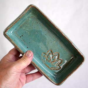 Può includere: Un vassoio rettangolare in ceramica con una smaltatura lucida color verde acqua e una texture maculata. I bordi sono rifiniti in una tonalità marrone a contrasto. Un motivo a fiore di loto in rilievo è centrato sul fondo del vassoio.
