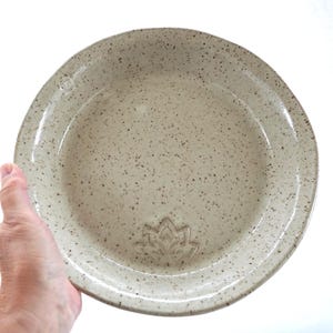 Puede incluir: Un plato de cerámica moteada de color blanquecino con un diseño de flor de loto. El plato tiene un borde ligeramente elevado y una forma de cuenco poco profunda. El plato está sostenido por una mano sobre un fondo blanco.